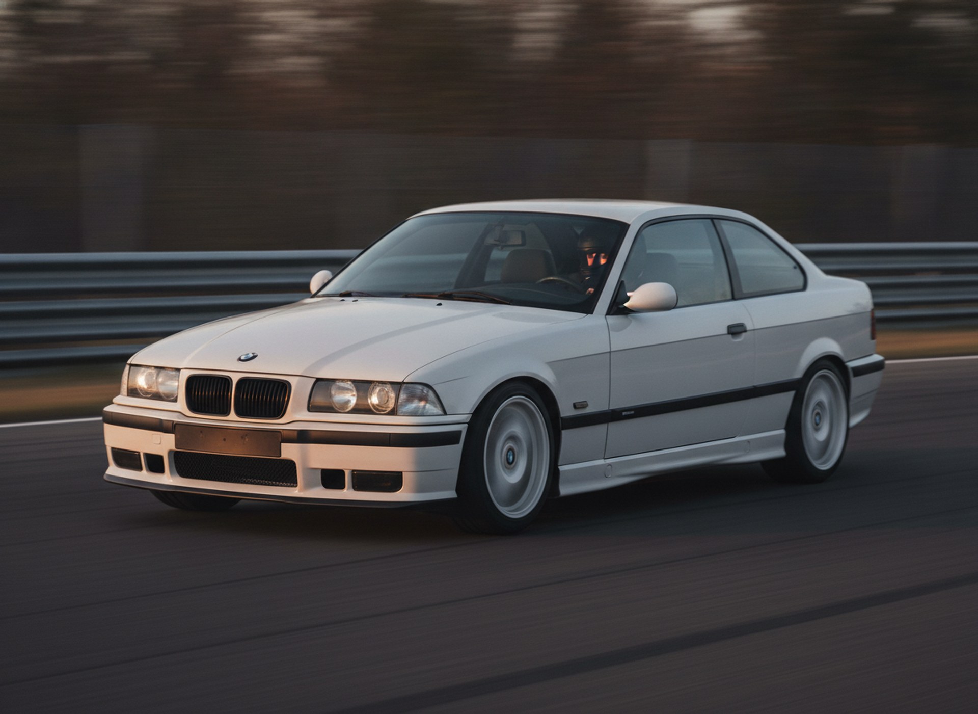 White BMW E36 3-Series coupe being driven hard through a corner on a race circuit, driver in a plain black helmet
