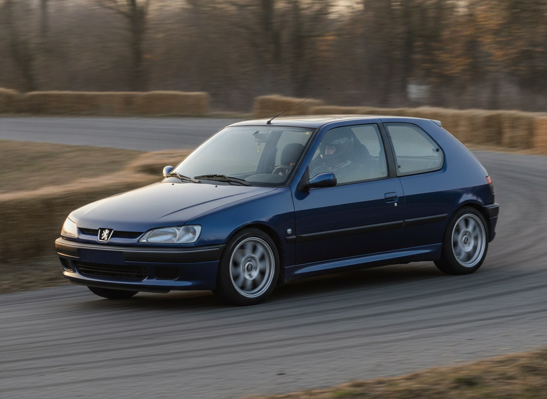 Blue Peugeot 306 GTI-6 being driven hard through a corner on a rally stage, driver in a plain black helmet