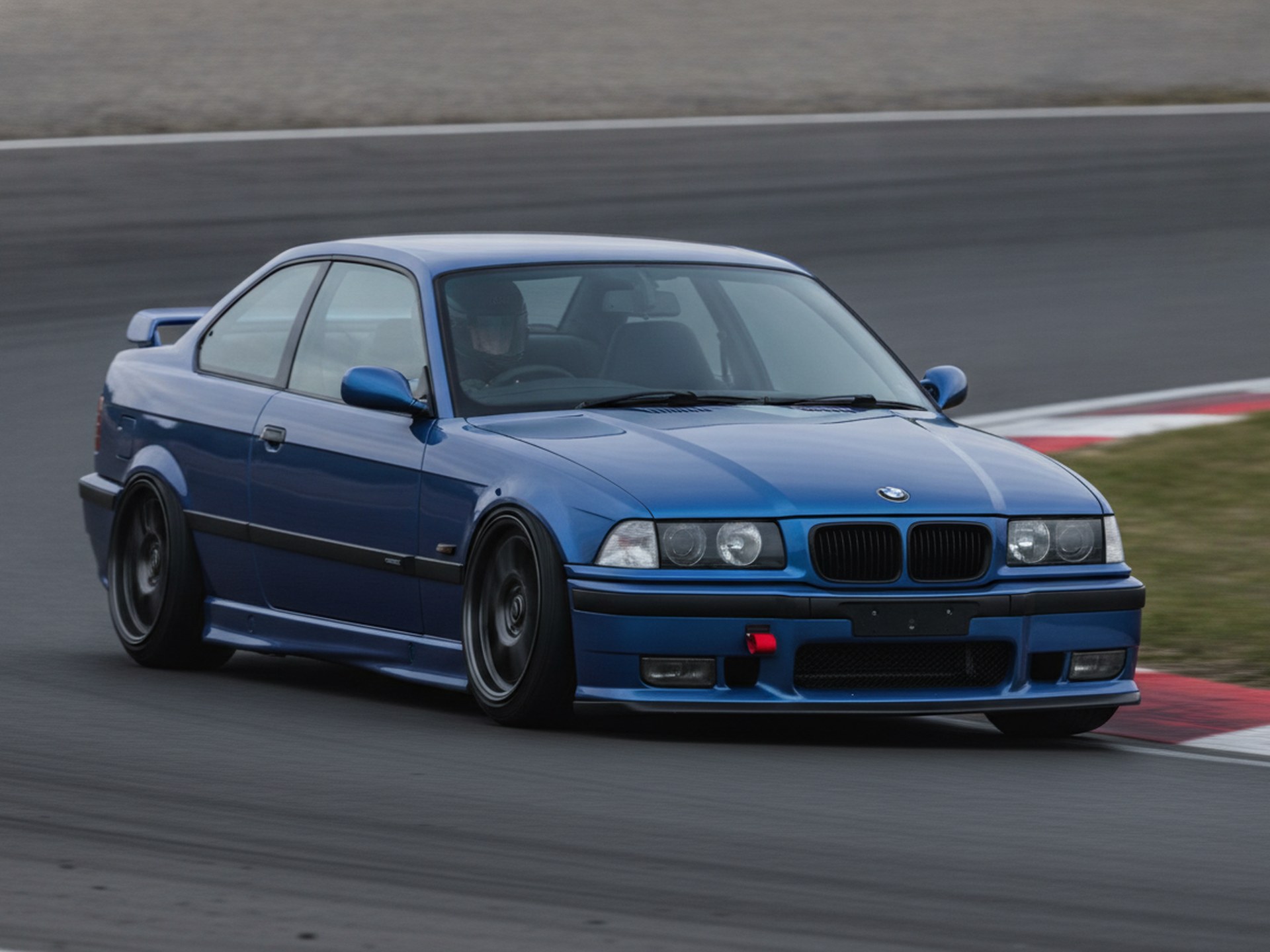 White BMW E36 3-Series coupe being driven hard through a corner on a race circuit, driver in a plain black helmet