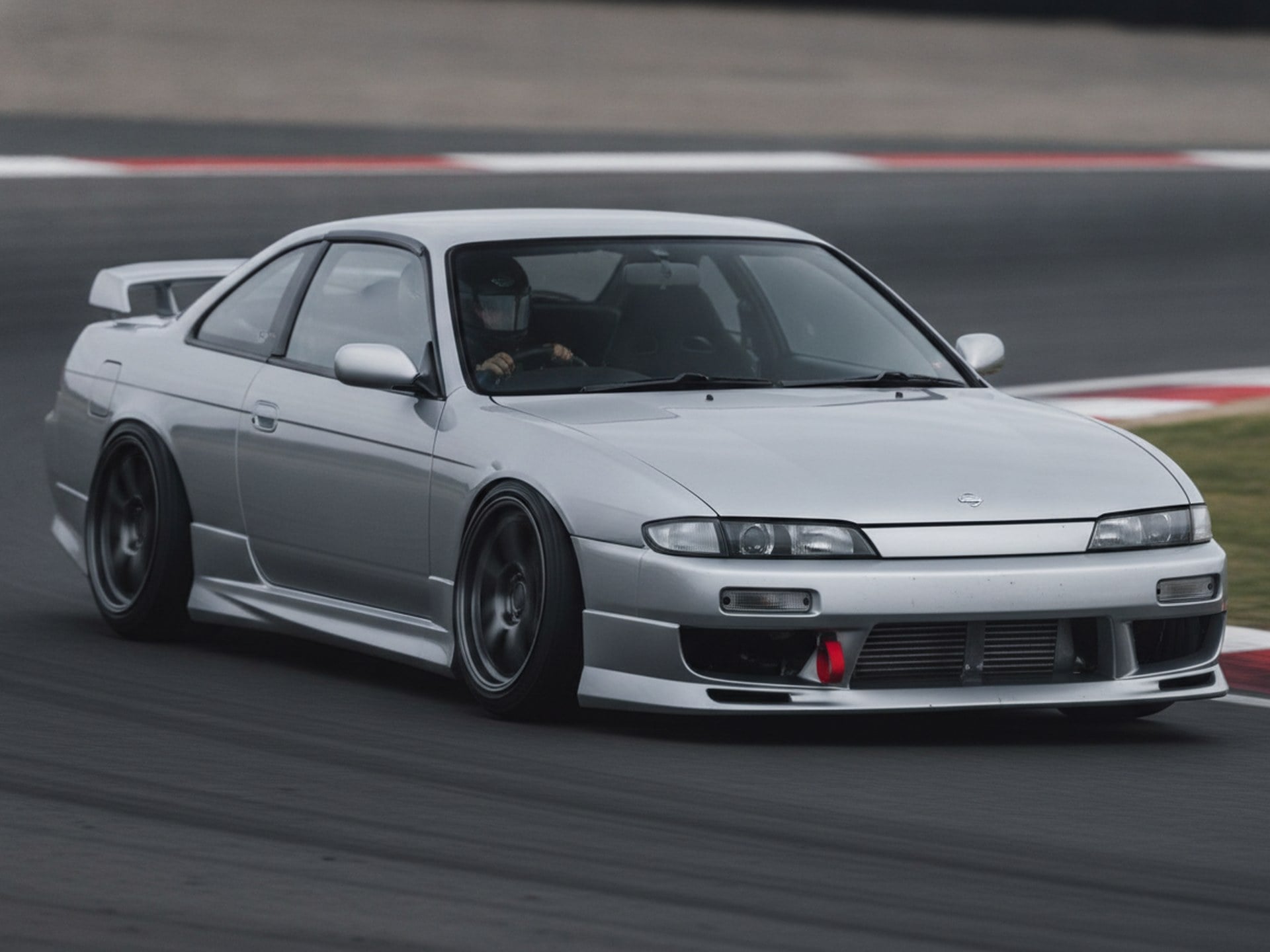 Silver Nissan 200SX S14 being driven hard through a corner on a tarmac rally stage, driver in a plain black helmet
