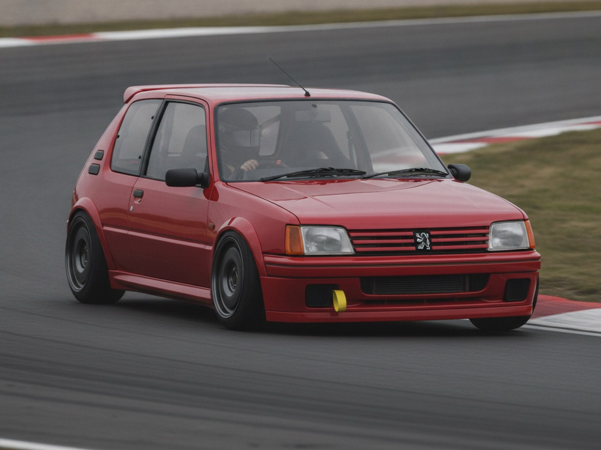 Peugeot 205 hot hatch cornering hard on a race circuit, driver in a plain black full-face helmet, big brake kit guide