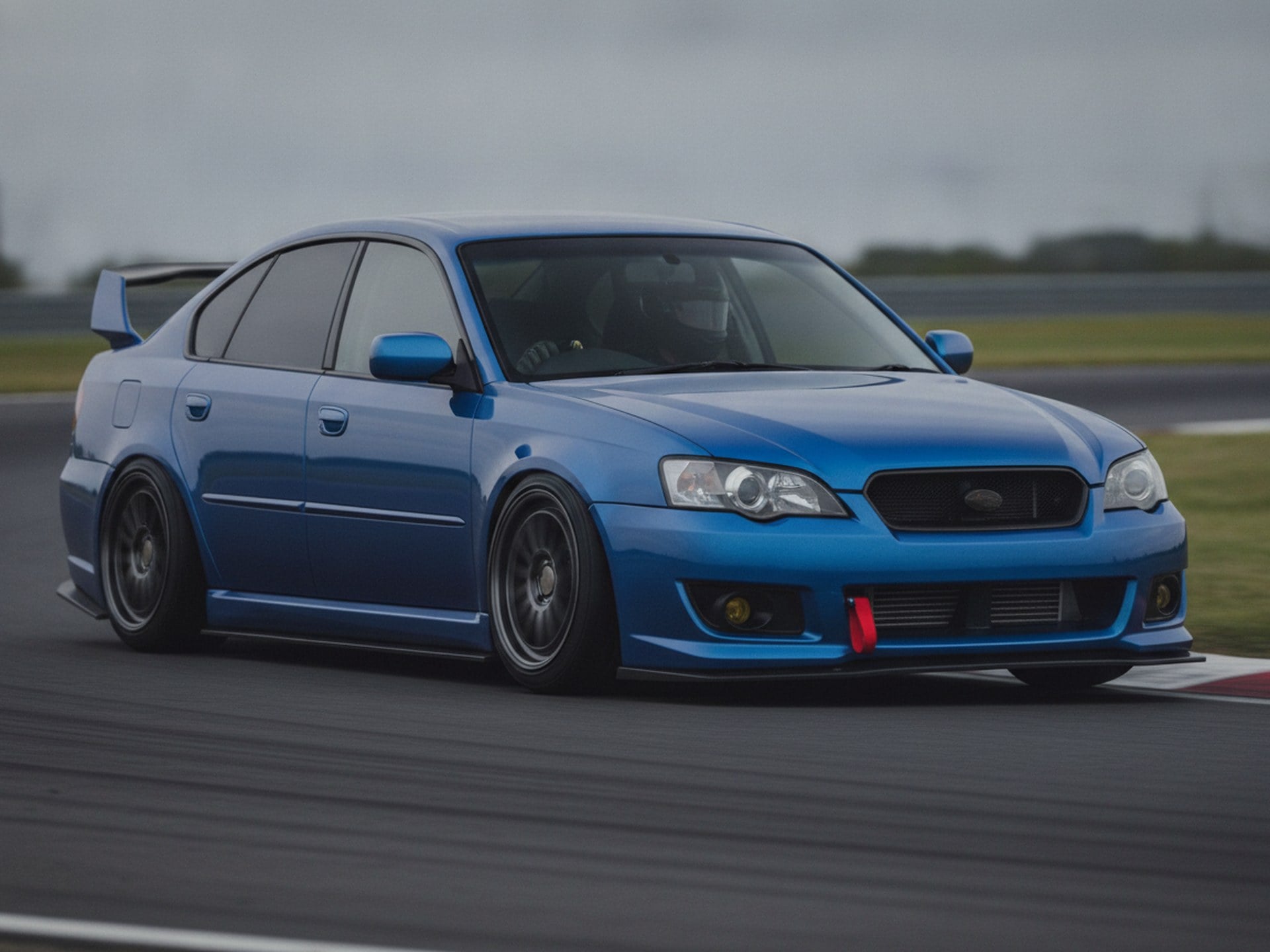 Blue Subaru Legacy saloon on a wet race circuit, driver in a plain black helmet, big brake kit guide